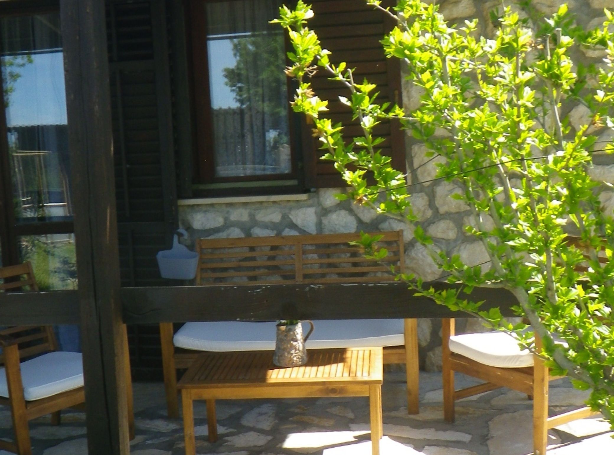 A wooden bench with a cushion, a small table, and a potted plant in a stone-walled outdoor area.