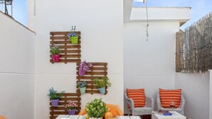 Un patio con una mesa preparada para el desayuno, que incluye zumo de naranja, bollería y una taza de café. Hay sillas de mimbre con cojines naranja y una pequeña mesa con una copa de vino tinto.