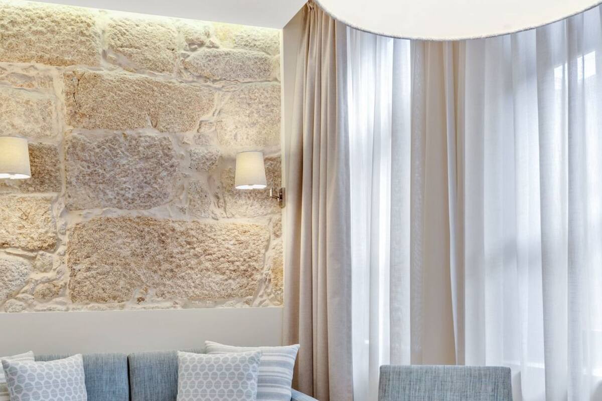 Un salon moderne avec un mur en pierre, un canapé bleu agrémenté de coussins à motifs, une table ronde sur laquelle reposent une carafe en cristal et des verres, et des rideaux légers.