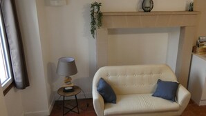 Een woonkamer met een beige bank, een klein bijzettafeltje met een lamp, een open haard en een raam met gordijnen.