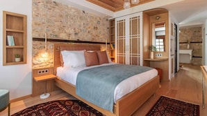 Une chambre avec un mur en pierre, un plafond en bois, un lit recouvert d’une couverture bleue, une table de chevet avec une lampe et une petite étagère avec des livres et une plante.