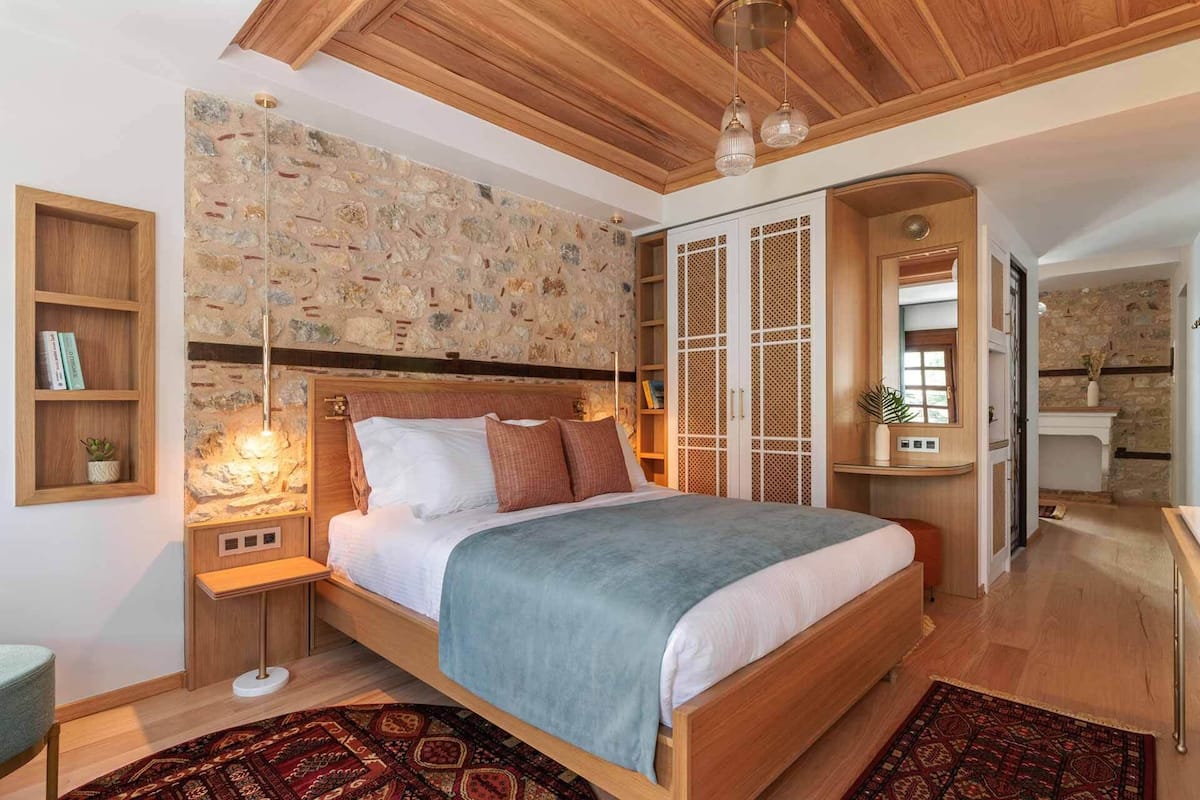 Un dormitorio con pared de piedra, techo de madera, una cama con manta azul, una mesita de noche con lámpara y un pequeño estante con libros y una planta.