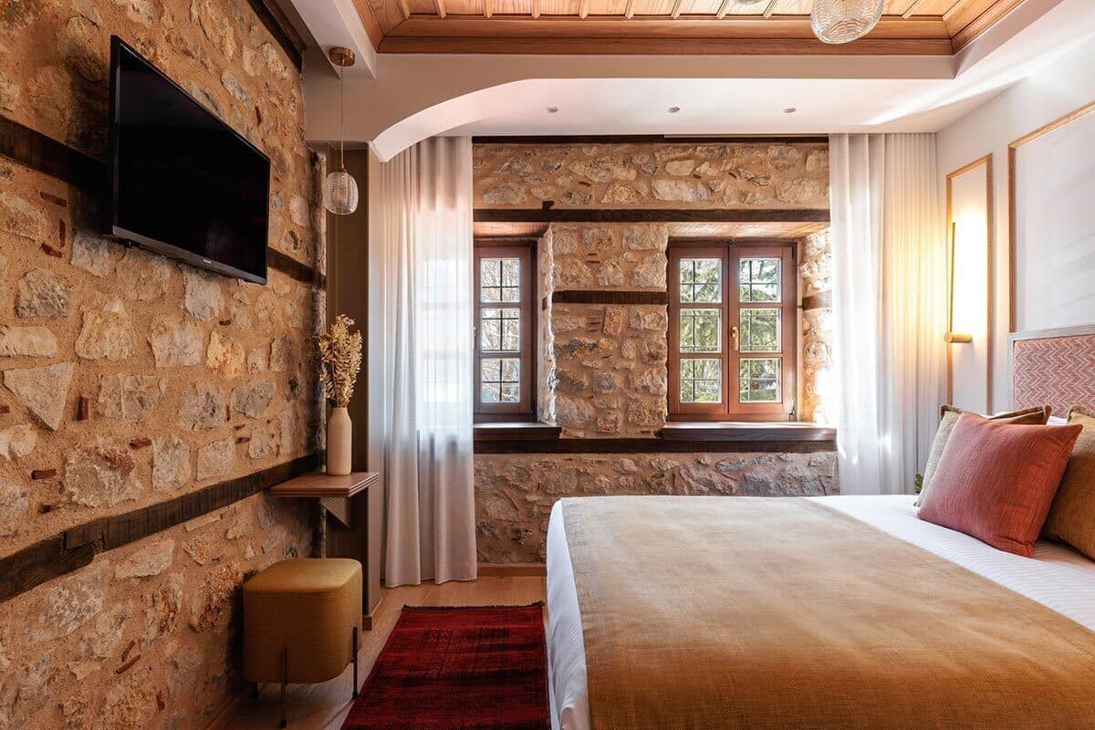 Un dormitorio con paredes de piedra, una cama grande, un televisor montado en la pared y una ventana con cortinas.