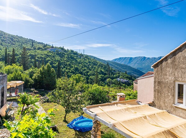 A rural landscape with a house featuring a sloped roof and stone walls, surrounded by lush greenery and mountains in the background. - Ranch House Zora (Dubrovnik)