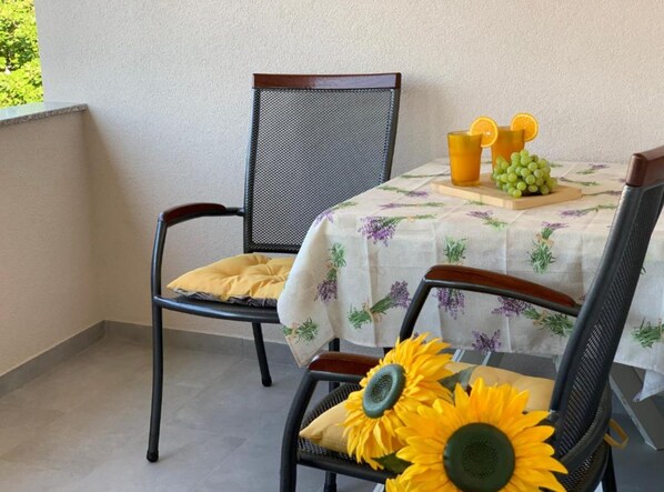 A balcony with two chairs and a table covered with a floral tablecloth. There are sunflowers on the table and a glass of orange juice with grapes.
