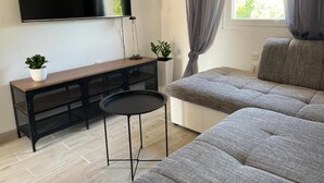 A modern living room with a grey sofa, a small black table, a TV mounted on the wall, and a window with curtains. - Apartments Rio (Biograd na Moru)