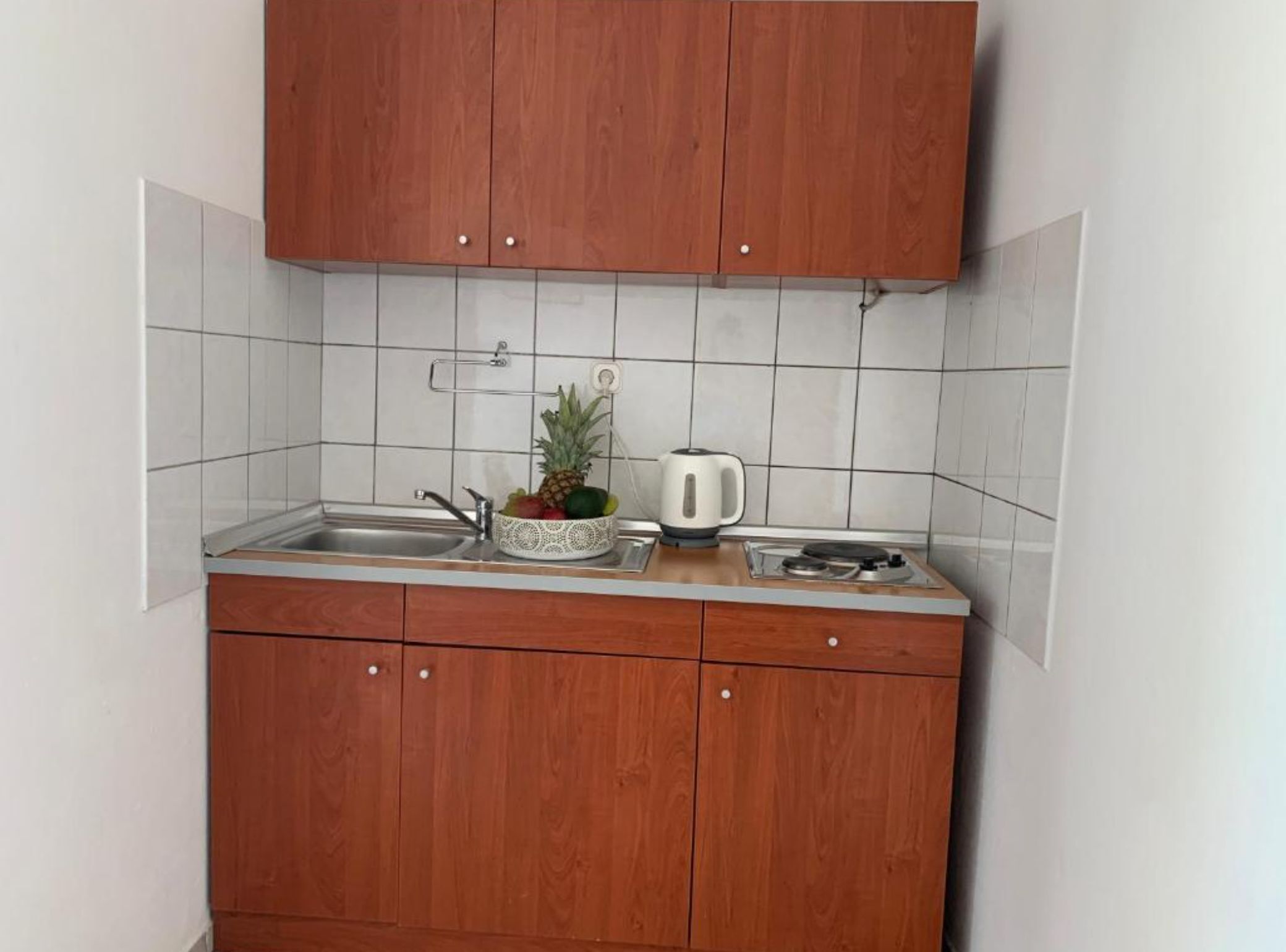 Une cuisine avec des meubles en bois, un crédence carrelée de couleur blanche, un évier, un panier de fruits contenant un ananas et une bouilloire.