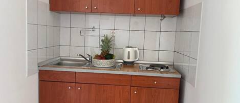 Une cuisine avec des meubles en bois, un crédence carrelée de couleur blanche, un évier, un panier de fruits contenant un ananas et une bouilloire.