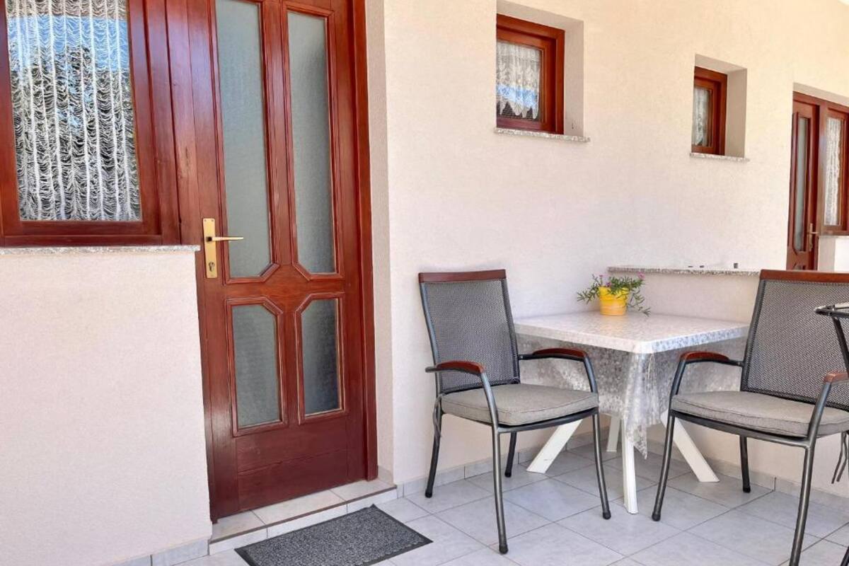 A room with a wooden door, a tiled floor, a white table with four chairs, and a small window with curtains.