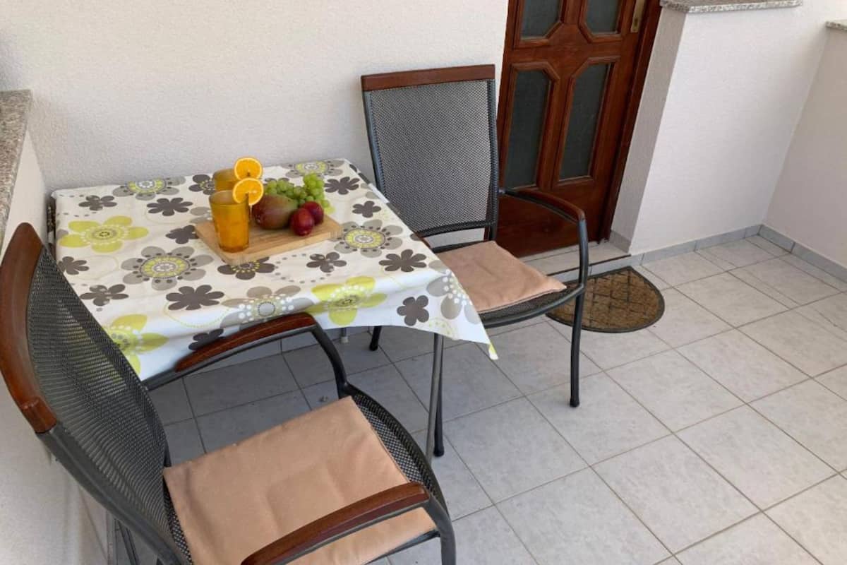A small outdoor dining area with a table covered by a floral tablecloth, two chairs, and a fruit basket with oranges and grapes.