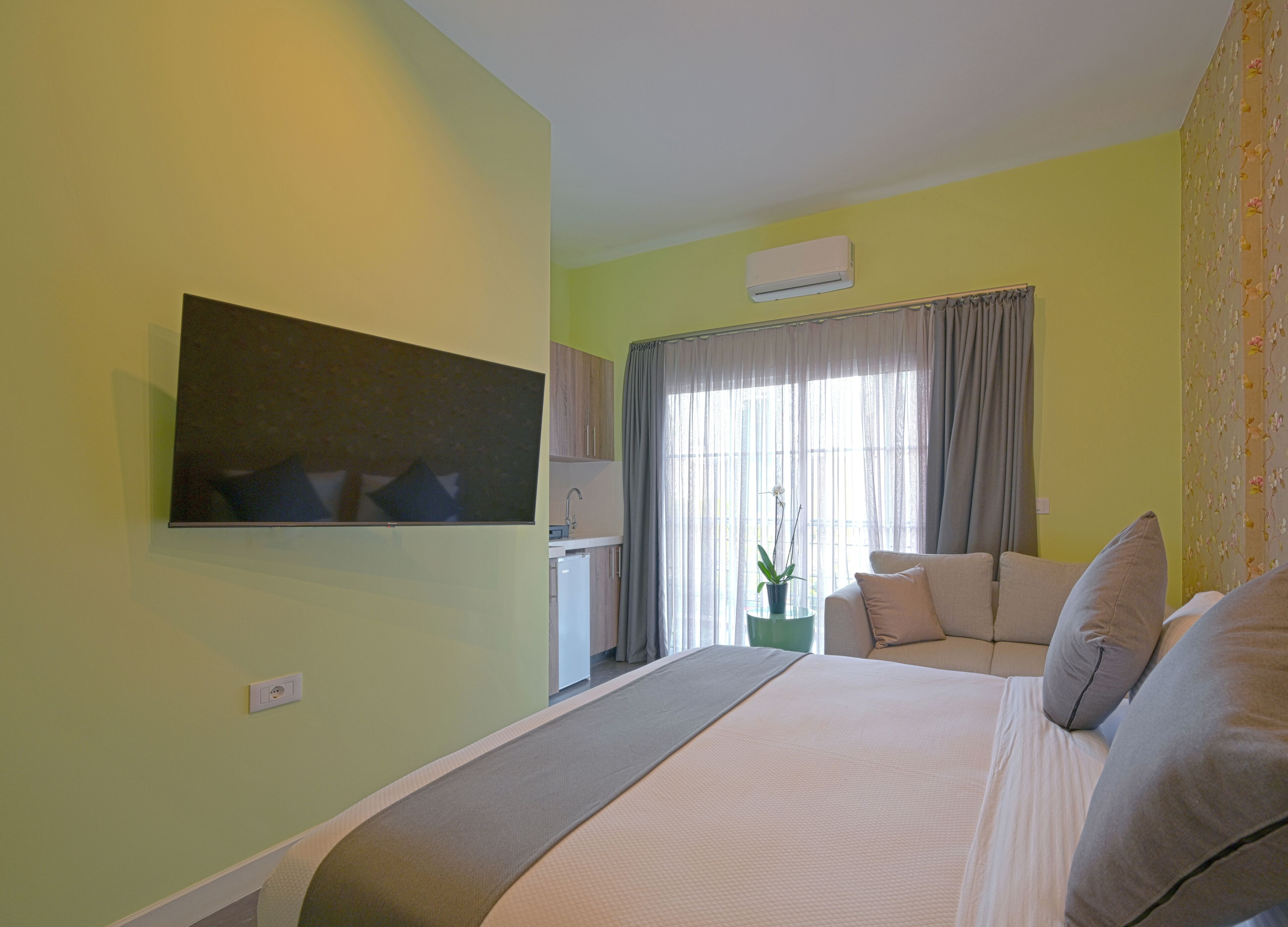 A hotel room with a bed, a TV mounted on the wall, a window with curtains, a small table with a plant, and a kitchenette area.