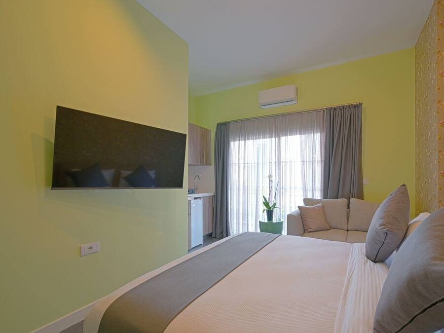 A hotel room with a bed, a TV mounted on the wall, a window with curtains, a small table with a plant, and a kitchenette area.