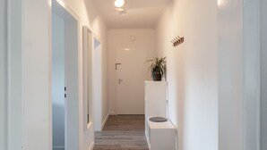 A hallway with a wooden floor, white walls, and a ceiling light. There is a door at the end of the hallway and a small plant on a white shelf. - Ferienwohnung Flut, Modern & Liebevoll Eingerichtet, Wlan, Smart TV (Wilhelmshaven)