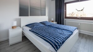 A modern bedroom with a bed, nightstands, a window with curtains, and a radiator. - Ferienwohnung Flut, Modern & Liebevoll Eingerichtet, Wlan, Smart TV (Wilhelmshaven)