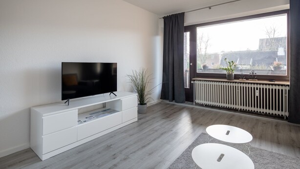 A modern living room with a flat-screen TV on a white console, a radiator, a large window with curtains, and two white coffee tables. - Ferienwohnung Flut, Modern & Liebevoll Eingerichtet, Wlan, Smart TV (Wilhelmshaven)