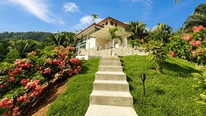 Une maison à deux étages avec un escalier en pierre menant à un balcon, le tout entouré d’une végétation luxuriante et de plantes en fleurs.