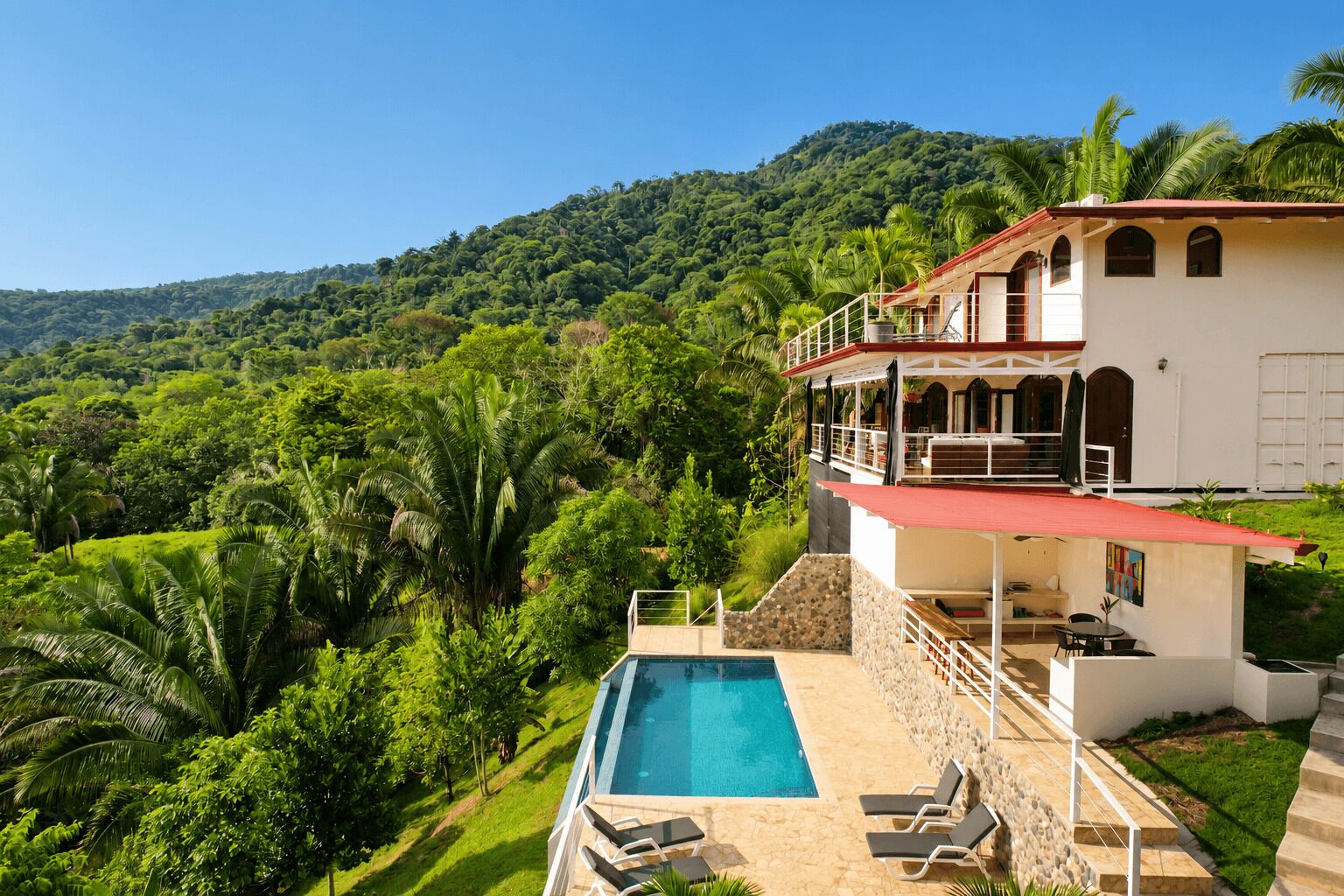 Um edifício de dois andares com piscina, cercado por vegetação exuberante e colinas.