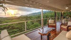 Une terrasse en bois avec des sièges rembourrés blancs et une table en bois, offrant une vue imprenable sur un paysage verdoyant et luxuriant, avec en toile de fond le coucher de soleil.