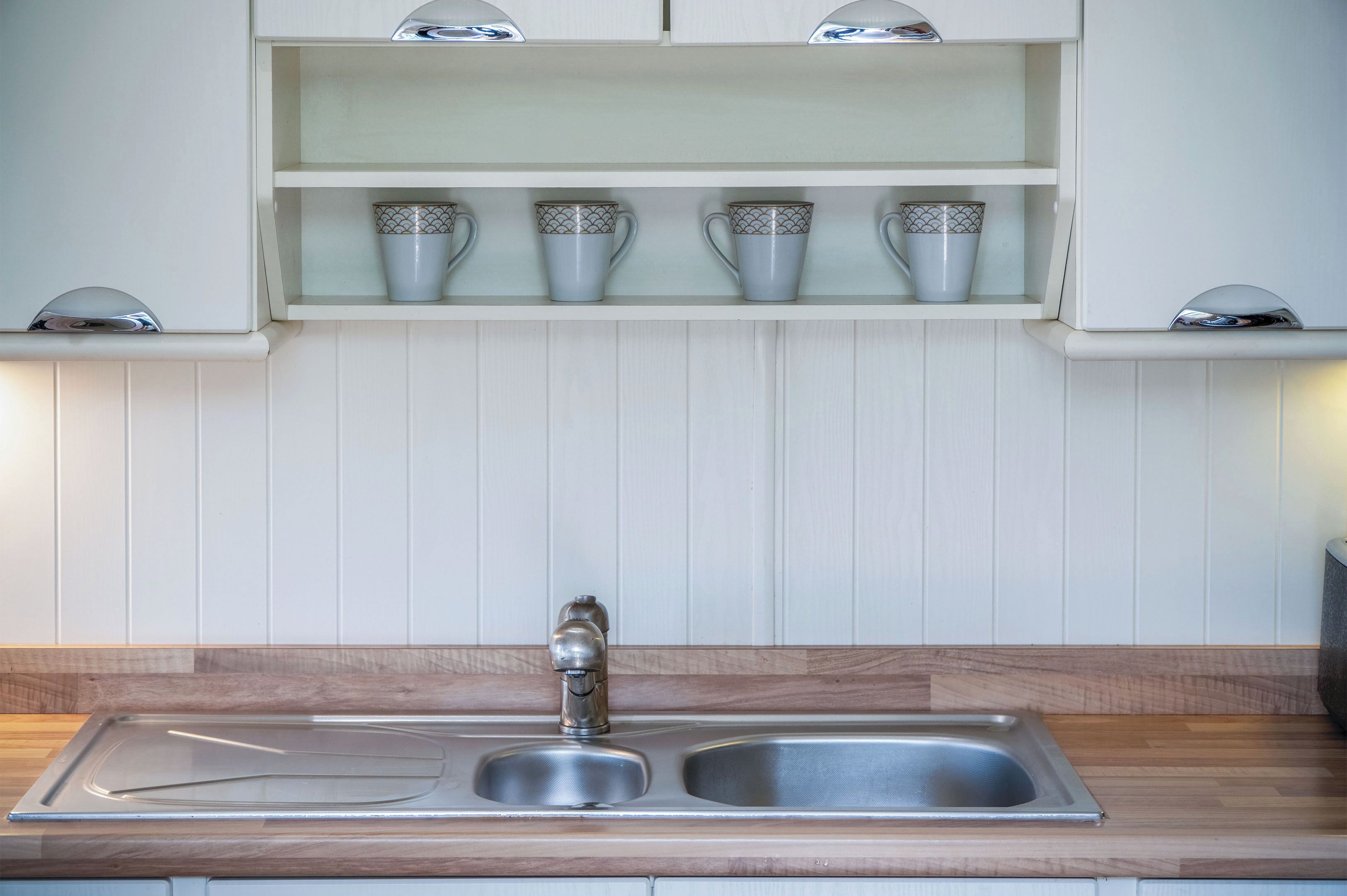 A kitchen with a double sink, white cabinets, and a shelf with four cups.