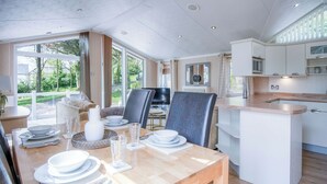 Un espace de repas moderne avec une table en bois disposée pour quatre personnes, une cuisine avec des meubles blancs et de grandes fenêtres offrant une vue sur l’extérieur.