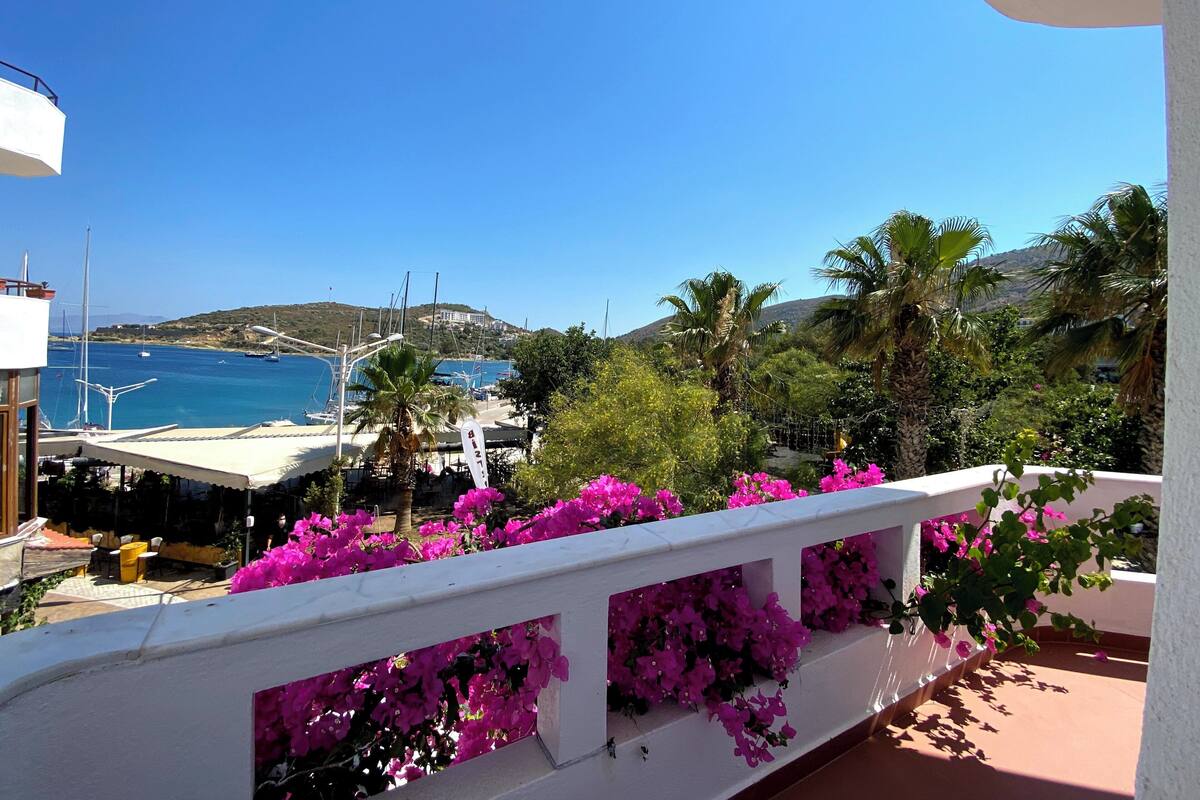 A balcony with white railings and pink flowers, overlooking a marina with boats and a clear blue sky.