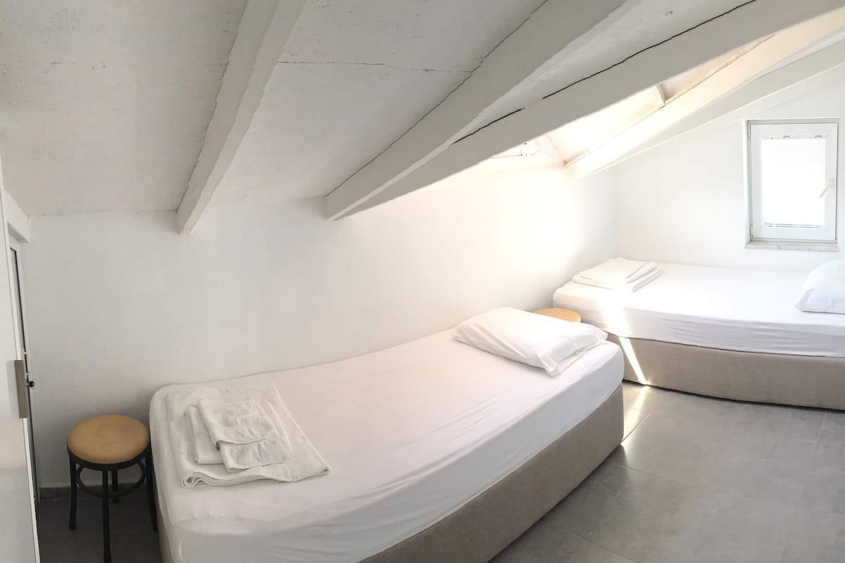 A compact bedroom with two beds, a small stool, and a window.