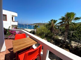 Un balcón con mesa de madera y sillas rojas, con vista a un puerto deportivo con veleros y un paisaje costero.