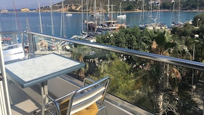 Un balcon avec une table et des chaises donnant sur une marina où sont amarrés des voiliers, et sur lequel on aperçoit, au loin, un bâtiment situé sur une colline.