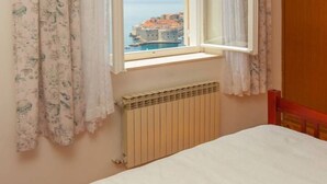 Un dormitorio con cama, ventana con cortinas, radiador y vistas a un pueblo costero.
