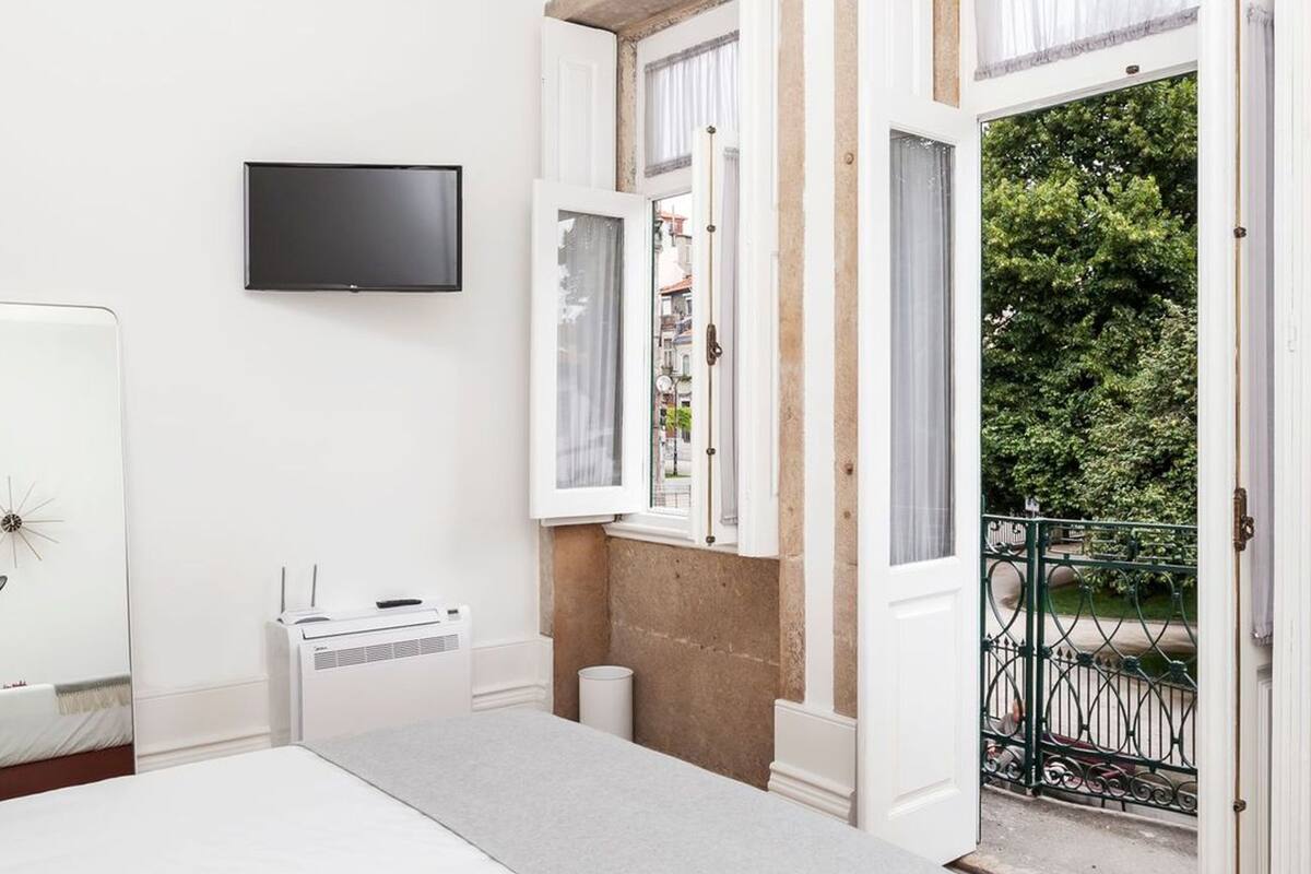 Een slaapkamer met een bed, een televisie, een verwarming en een raam met uitzicht op een balkon en groen.