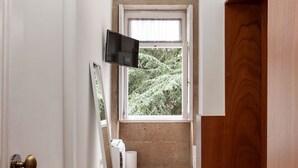A narrow hallway with a wooden floor, a window with a view of trees, a wall-mounted TV, and a door with a brass handle. - Passeio de São Lázaro nº 5 (Porto)