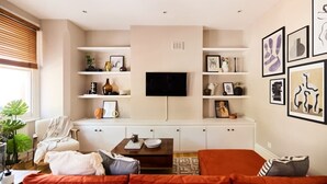 A modern living room with a sofa, coffee table, and TV. Shelves with decorative items and framed pictures are on the walls. - The Waterlow Park Wonder - Trendy 2bdr With Study Room and Patio (London)