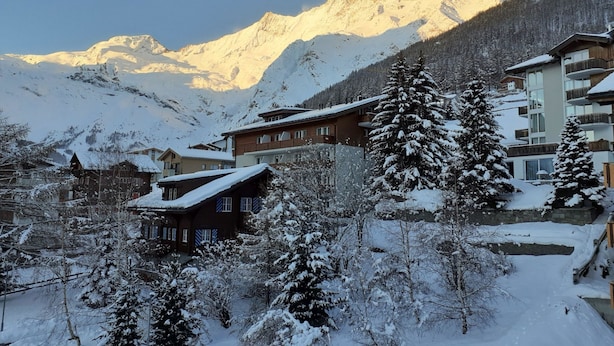 Un village de montagne enneigé, avec ses maisons traditionnelles en bois et ses bâtiments modernes, le tout dominé par un impressionnant paysage montagneux.