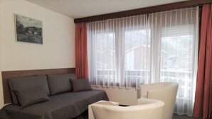 Habitación de hotel con cama, sofá, dos sillones y un ventanal con cortinas traslúcidas.