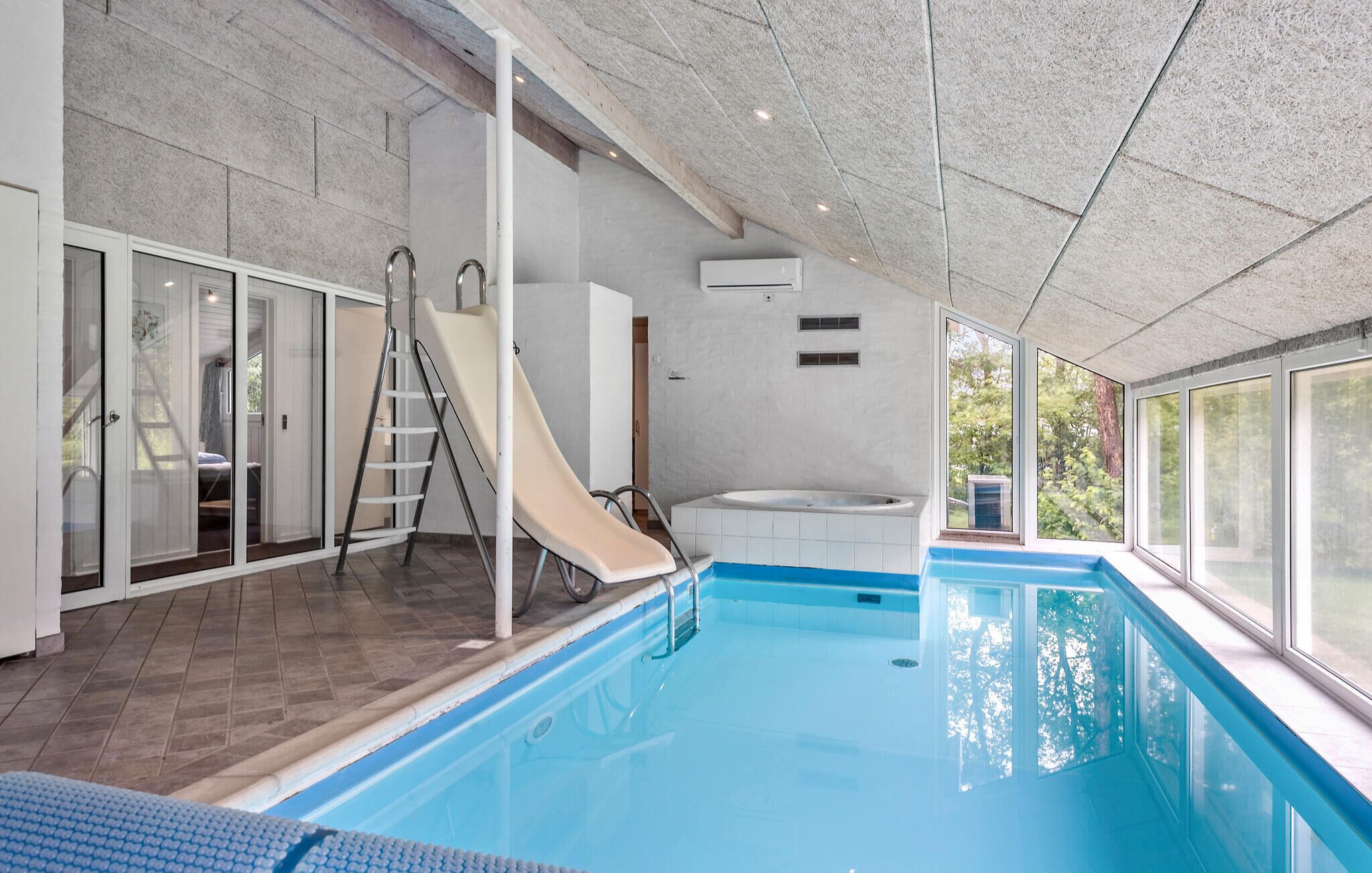 An indoor swimming pool with a slide, a hot tub, and a view of trees through large windows.