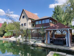 Una casa de dos pisos amb teulada de rajola vermella, terrassa de fusta i una petita bassa.