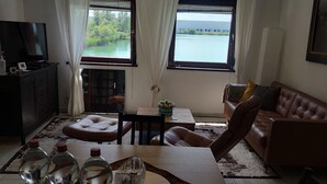 Ein Wohnzimmer mit einer Couch, einem Fernseher, einem Esstisch mit Wasserflaschen und Blick auf ein Gewässer durch große Fenster.