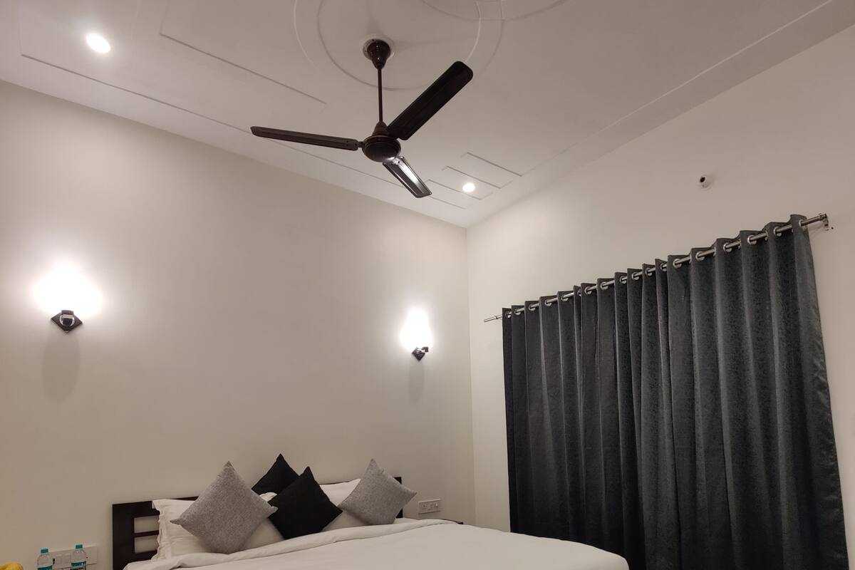 A bedroom with a bed, a ceiling fan, two wall-mounted lights, a dark curtain, and a small table with a water bottle.