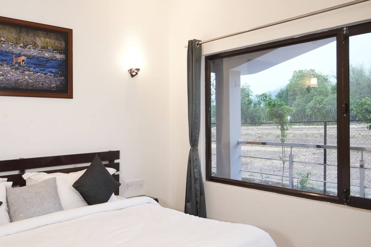 A hotel room with a bed, pillows, a painting of a tiger by a river, and a window with a view of a fenced area and trees.