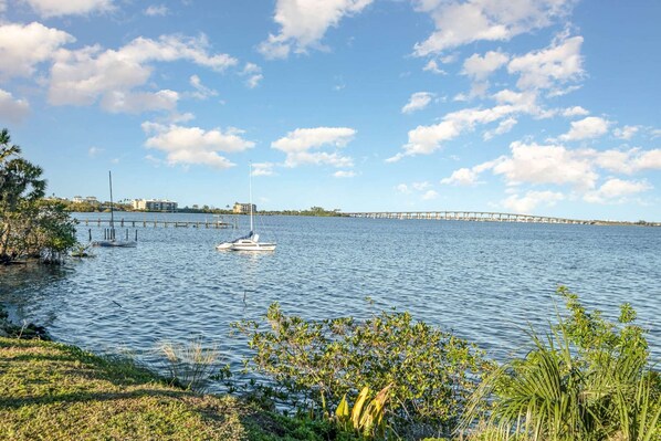 A marina with a sailboat docked, a bridge in the background, and a grassy shoreline with vegetation.