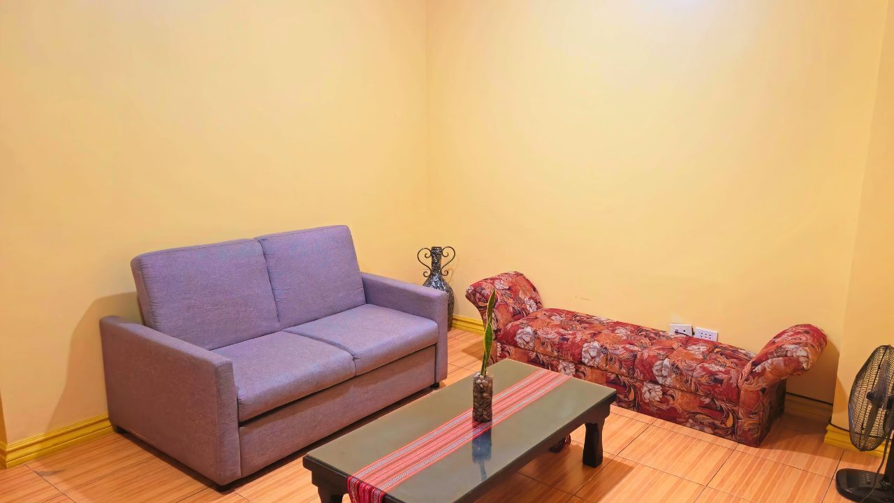 A living room with a purple sofa, a floral sofa, a coffee table with a vase, and a standing fan.