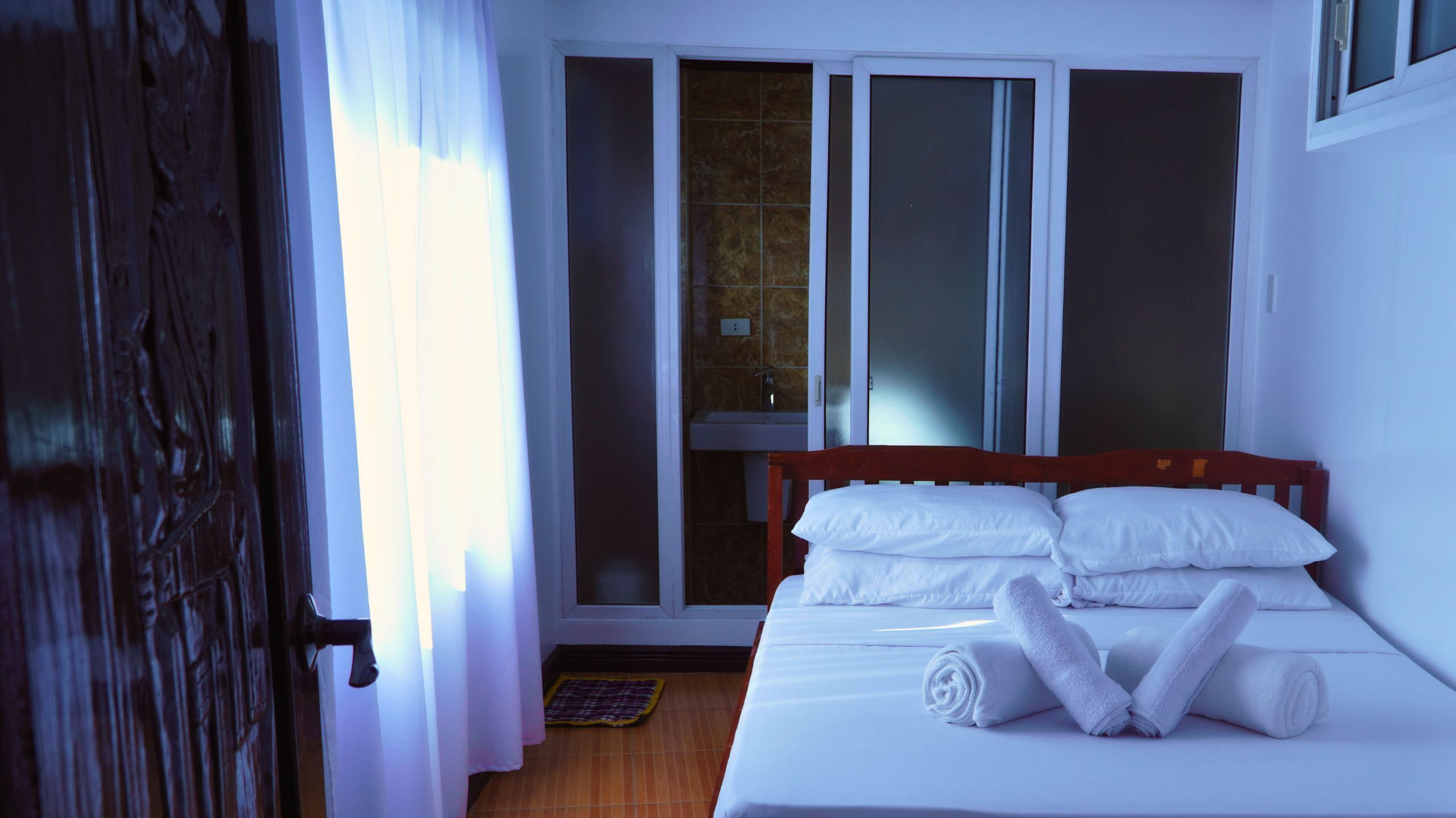 A hotel room with a bed, white linens, a wooden headboard, a window with curtains, and a bathroom with a sink and mirror.