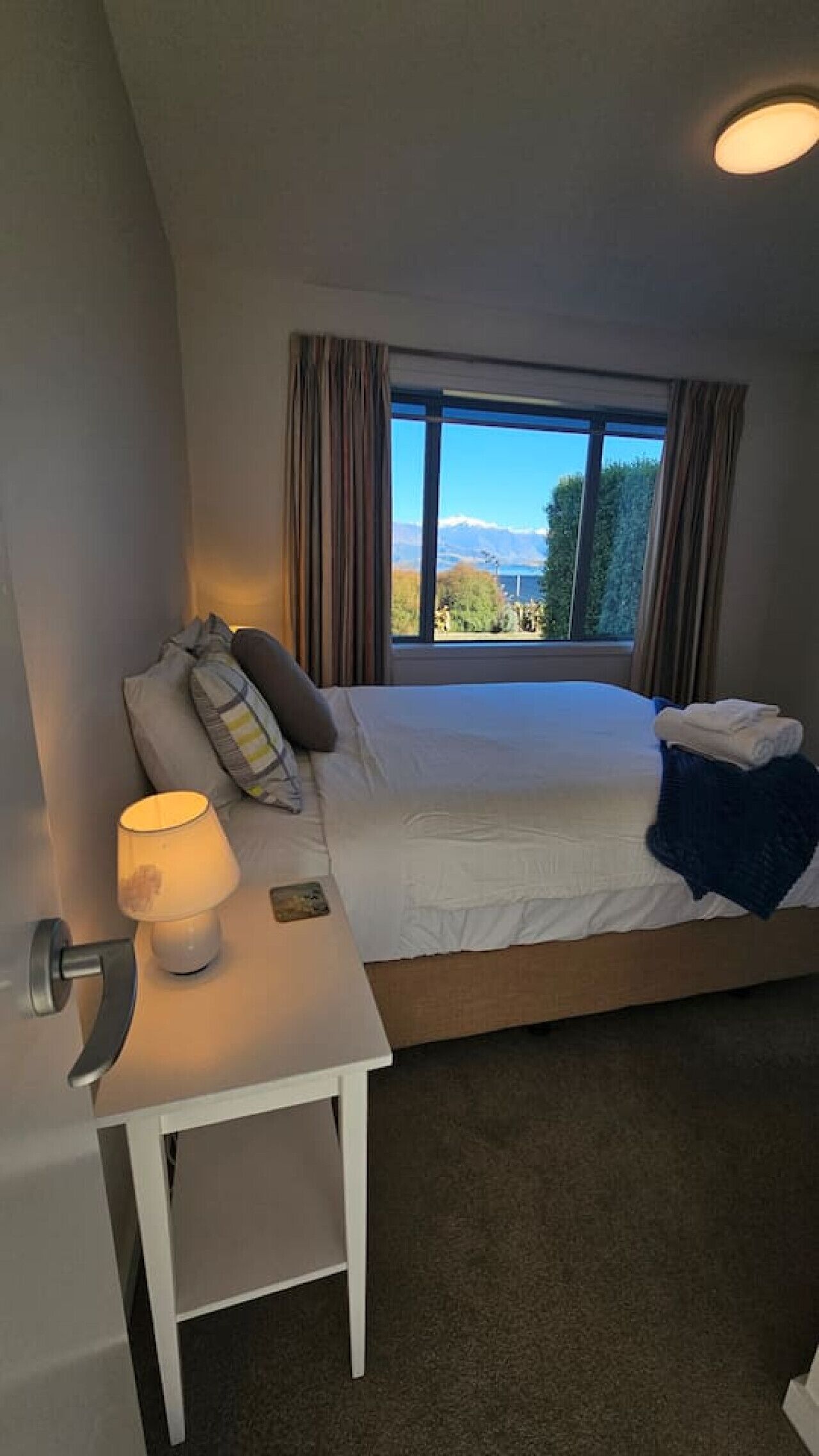 Un dormitorio con una cama, una mesita de noche con una lámpara, vistas a las montañas a través de una ventana y una puerta.