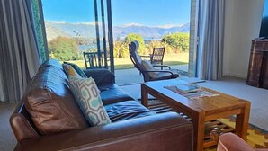 A living room with a brown leather sofa, a wooden coffee table, and two chairs. There is a large window with curtains, offering a view of mountains and greenery.