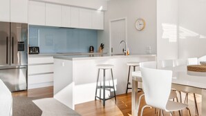 A modern kitchen with white cabinetry, a stainless steel refrigerator, a blue backsplash, and a dining area with white chairs and a table. - Moments from That Wanaka Tree - only a 5-minute walk to That Wanaka Tree (Wānaka)