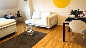 Un salon moderne avec un canapé blanc, une petite table basse, une table à manger avec une chaise, et un élément de décoration jaune sur le mur.