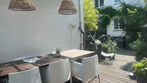 Une terrasse extérieure avec une table en bois et des chaises blanches, entourée de plantes en pot et agrémentée d’un luminaire suspendu.