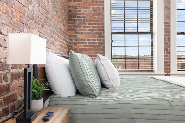 Quarto com parede de tijolos, janela com persianas, cama com manta listrada, abajur, controle remoto e planta em vaso.