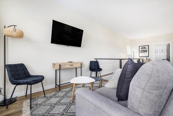A modern living room with a grey sofa, two black chairs, a white coffee table, a TV on the wall, a desk with a chair, and a patterned rug.