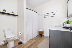 Een moderne badkamer met toilet, douchegordijn, een klein krukje en een wastafelgedeelte met een plant.